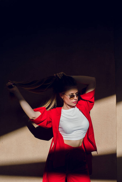 Πορτρέτο μιας νεαρής ξανθιάς με κόκκινο κοστούμι που ποζάρει. Σκληρές σκιές από τον ήλιο πέφτουν πάνω της. Κορίτσι κρατά τα μαλλιά χέρι. - Φωτογραφία, εικόνα