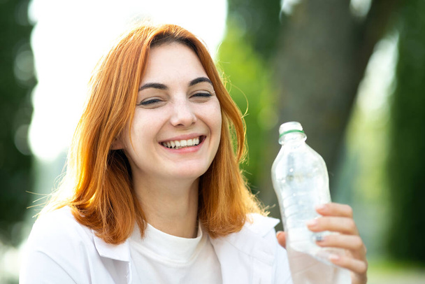 Junge durstige rothaarige Frau trinkt Wasser aus einer Flasche im Sommerpark. - Foto, Bild