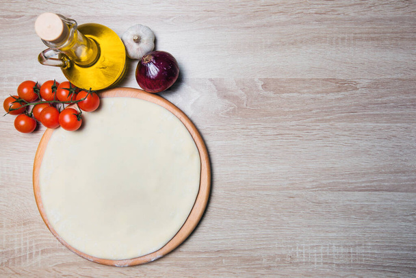 Dough basis for the pizza on a wooden board and cherry tomatoes, olive oil, onions, garlic - Foto, Imagen