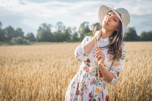 joven, emocional, chica bonita en un vestido ligero con un sombrero de luz al atardecer en un trigo, campo de oro, sosteniendo espigas de trigo
  - Foto, imagen
