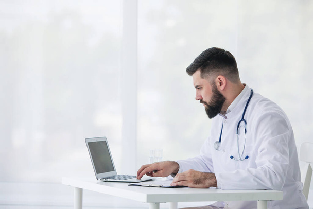 Dokter werkt met documenten en laptop in zijn medische kantoor - Foto, afbeelding