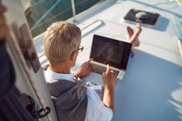 Senior man is working during the vacation on a sailboat - Foto, Imagen