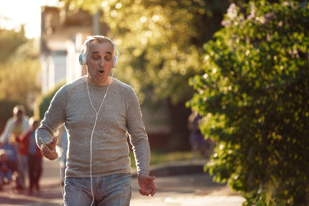 Senior man is walking along the sidewalk listening to music and singing a song - 写真・画像