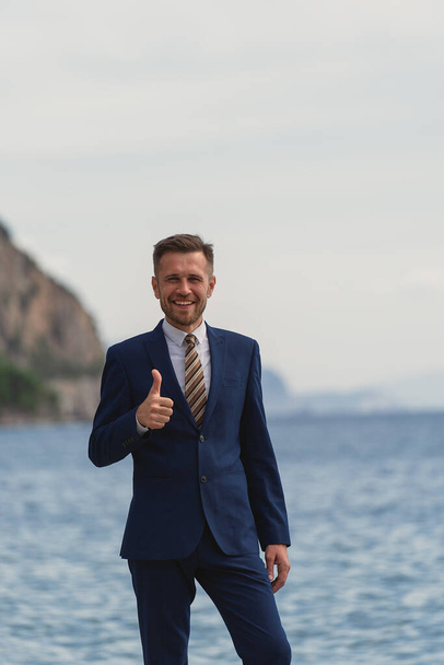 Businessman standing on the beach in a suit. Looks at camera and smiling broadly showing thumbs up gesture - Photo, Image