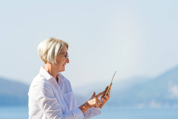 Mature woman in eyeglasses using tablet on background the sea and blue sky - Valokuva, kuva