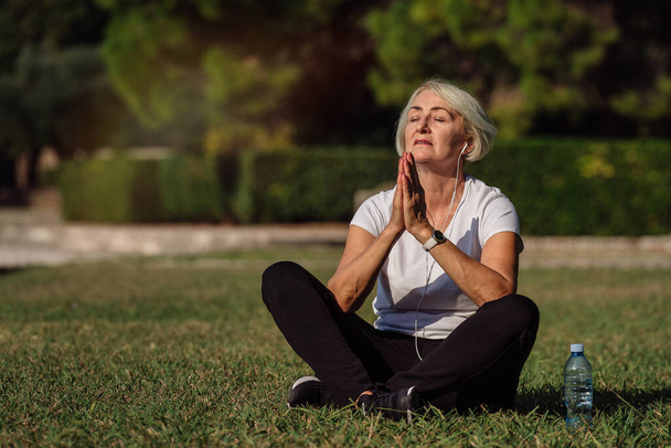 Mature woman sitting on the grass, resting after a run and listening to music - Foto, Imagem