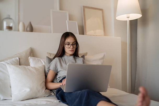                                Cute teenager girl doing homework lying on bed at home. Young pretty girl wearing glasses studying online with laptop, distance learning, self education     - Фото, изображение