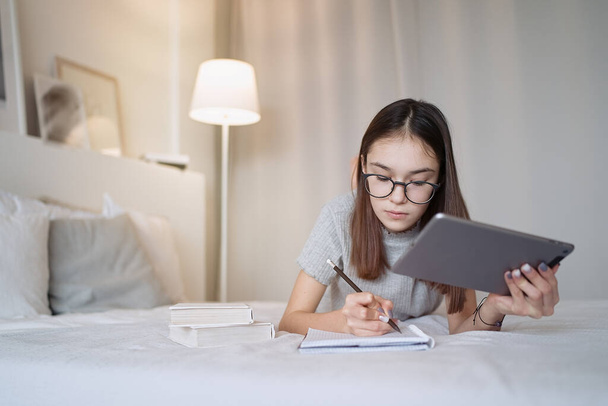                                Cute teenager girl using tablet lying on bed at home. Young pretty girl wearing glasses studying online, distance learning, self education - Fotografie, Obrázek
