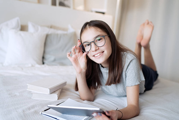                                Ragazza adolescente carina utilizzando tablet sdraiato sul letto a casa. Giovane bella ragazza con gli occhiali che studiano online, l'apprendimento a distanza, l'autoeducazione - Foto, immagini
