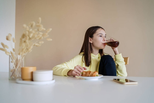                                Cute teen girl eating breakfast in kitchen while using on her smart phone   - Foto, Imagen