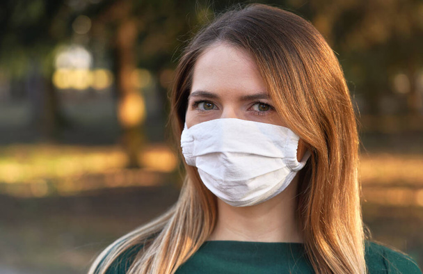 Young woman wearing white cotton virus mouth nose mask, blurred park and trees background, closeup face portrait - Photo, Image