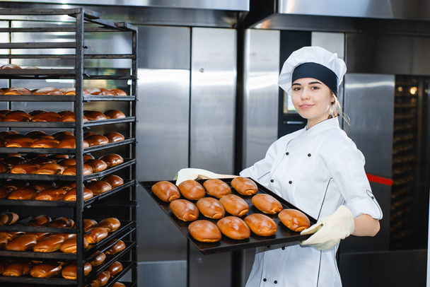 Young girl baker holds a baking tray with hot pastries on the background of an industrial oven in a bakery. - Foto, immagini