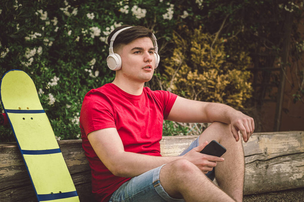 Teen Boy está sentado descansando con Longboard - Jóvenes adultos patinaje al aire libre - Un joven está sentado con un longboard, sosteniendo un teléfono en sus manos, escuchando música con auriculares
 - Foto, Imagen