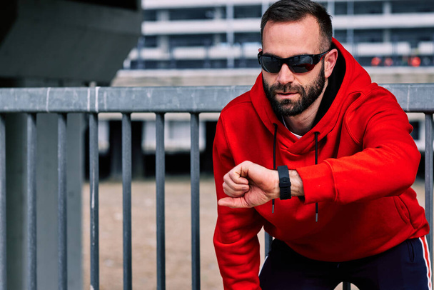 Attractive bearded runner in tracksuit resting and looking at smart watch his pulse. - Photo, Image