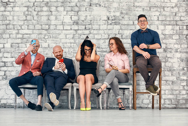 Group of smiling young business people sitting in line using mobile phones and other devices over white brick background - Fotó, kép