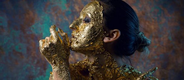 Mädchen mit Maske auf dem Gesicht aus Blattgold. Düsteres Studioporträt einer Brünetten auf abstraktem Hintergrund. - Foto, Bild