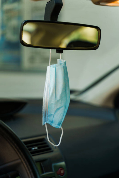 Medical protective face mask in car on the rearview mirror in day - Covid-19 pandemic protection on rear view in the vehicle - coronavirus epidemic new normal prevention concept - Photo, Image