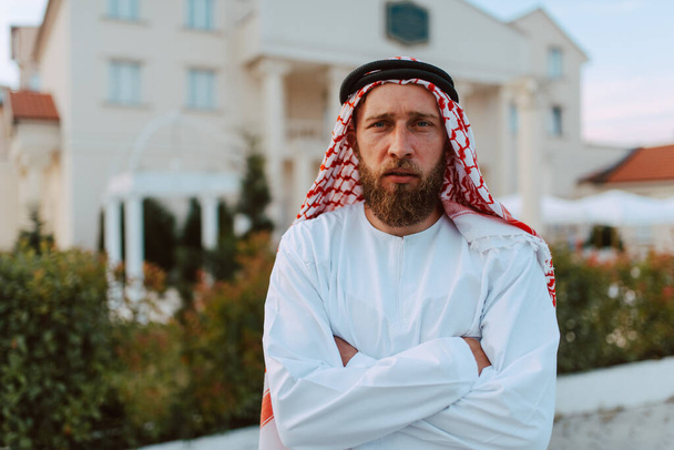 Portret van de knappe Midden-Oosterse Arabische moslim man in traditionele kleding die voor het witte mooie moderne huis staat - Foto, afbeelding