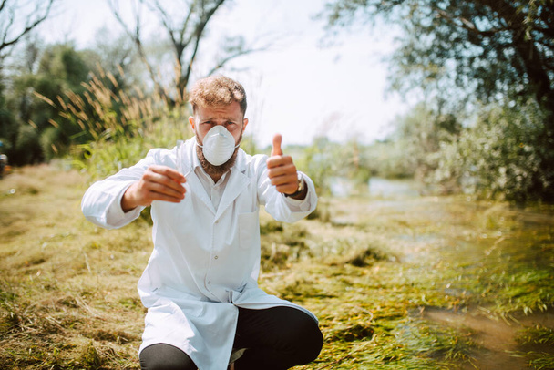 Мужчина, ученый-биолог и исследователь в защитном костюме, берёт пробы воды из загрязненной реки
. - Фото, изображение