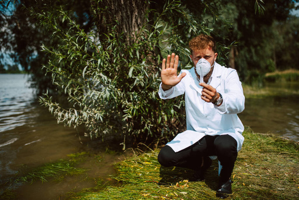 Mężczyzna kaukaski naukowiec biolog i badacz w kombinezonie ochronnym i masce pokazuje znak stopu i pobierania próbek wody z zanieczyszczonej rzeki. Kontrola wody - Zdjęcie, obraz