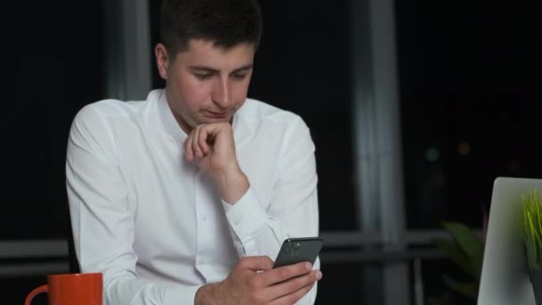 Cute young man with a Caucasian appearance lingered in the office at work until night. Sits on the phone in social networks and smiles - Filmagem, Vídeo