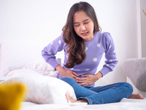 Mulheres asiáticas com menstruação e cólicas menstruais. uma menina sentada na dor em uma cama em casa. - Foto, Imagem