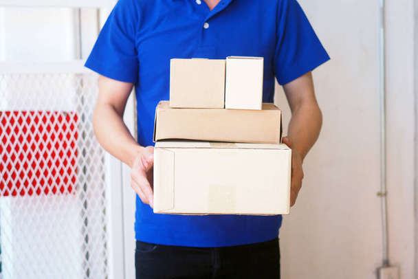 El remitente en el traje azul sostiene una caja marrón de pie frente a la casa del cliente. Envío gratuito para compras en línea y entrega urgente - Foto, Imagen