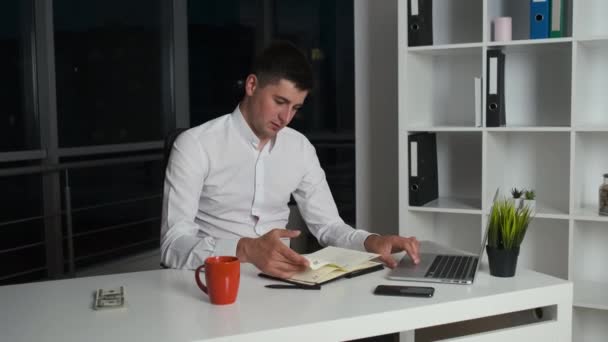 The 30-year-old director of the company sits in the evening office until late drinking coffee or tea and reading a book. Panorama of night city from the office - Video, Çekim