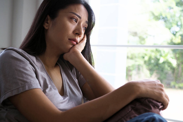 Азиатка, сидящая в доме и смотрящая в окно. Женщина смущена, разочарована, расстроена и расстроена - Фото, изображение