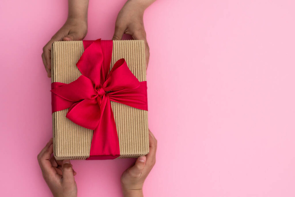 Mãos dando ou recebendo um presente em papel artesanal com uma fita rosa no fundo rosa pastel. Vista superior. Espaço de cópia
 - Foto, Imagem