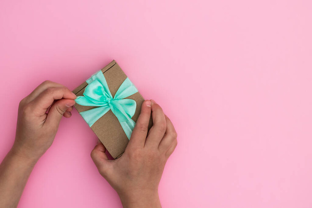 Handen geven of ontvangen van een geschenk in ambachtelijk papier met een muntlint op pastelroze achtergrond. Bovenaanzicht. Kopieerruimte - Foto, afbeelding