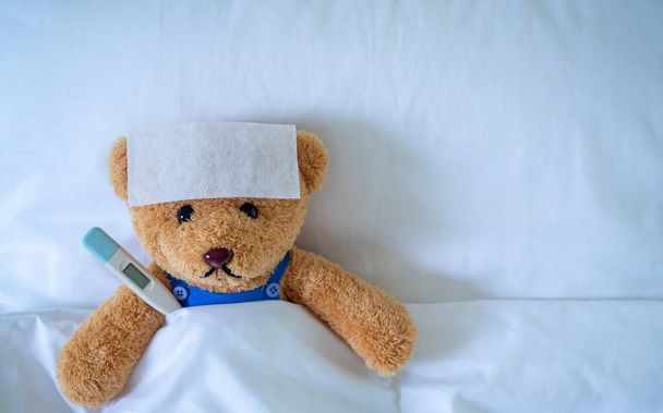 Brown teddy bear with fever reducing sheet and body temperature monitor. Used for health insurance or illness concepts - Photo, Image