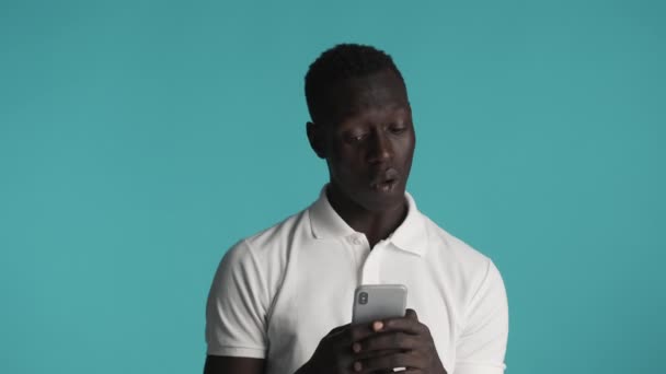 Mladý pohledný ležérní Afroameričan, který upřeně používá smartphone na barevném pozadí. Wow výraz - Záběry, video