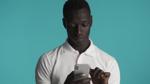 Młody, atrakcyjny Afroamerykanin używający smartfona na niebieskim tle. Wyrazy twarzy - Materiał filmowy, wideo