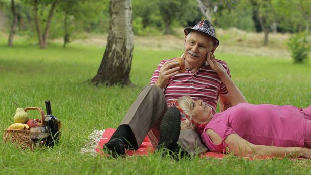 Piknik weekendowy w parku. Aktywny starszy stary kaukaski para siedzieć na koc i pić wino - Zdjęcie, obraz