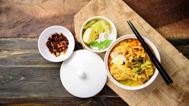 Северная тайская еда (Khao Soi), острый суп с лапшой из карри с курицей в миске, поедающей хрустящую яичную лапшу, маринованную горчицу, лук-шалот, лайм и молотый чили, жареный в масле - Фото, изображение