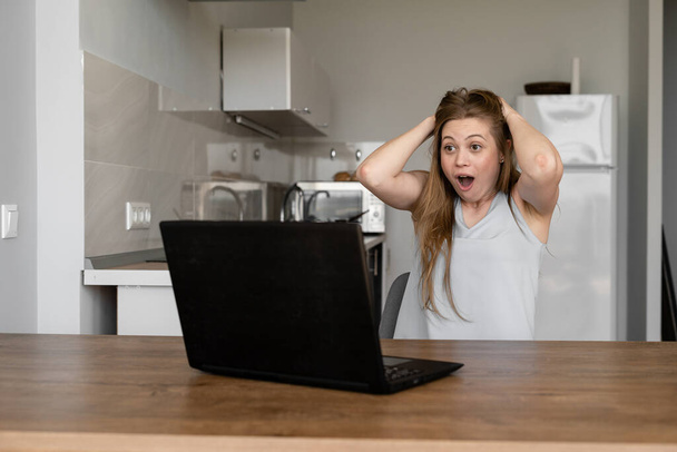mujer sorprendida sentada en casa en un portátil. Noticias inesperadas. Leer noticias en línea
. - Foto, imagen