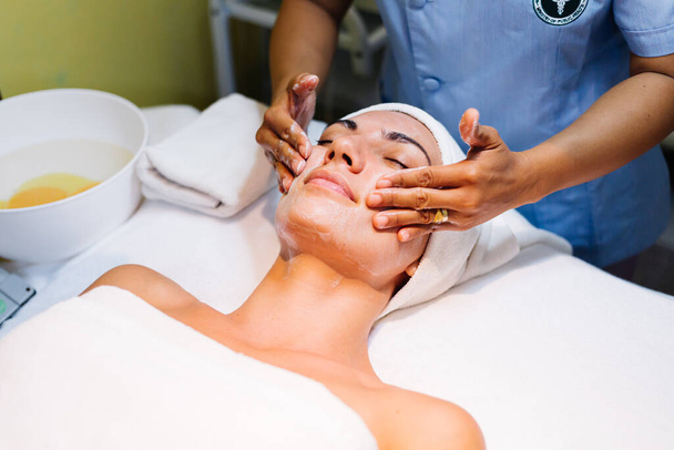 Junge Frau liegt während einer Verjüngungskur auf dem Kosmetiktisch. Kosmetikerinnen kümmern sich um jugendliche Haut an Hals und Gesicht und um Wohlbefinden. Reinigung der Hardware. - Foto, Bild