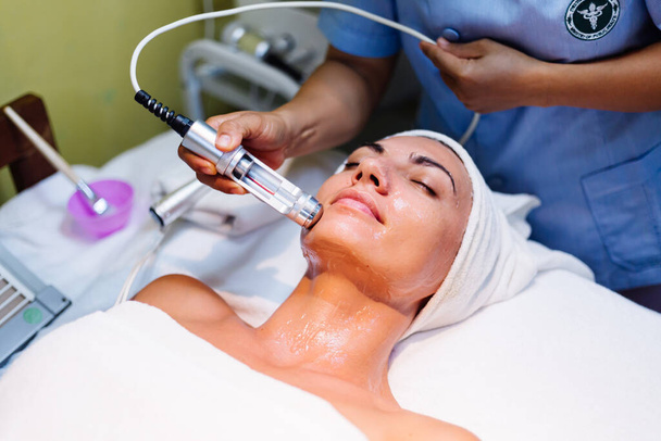 Молодая женщина лежит на косметологе во время процедуры омоложения. Косметолог заботится о коже шеи и лица молодой и оздоровительный. Аппаратная очистка лица. - Фото, изображение