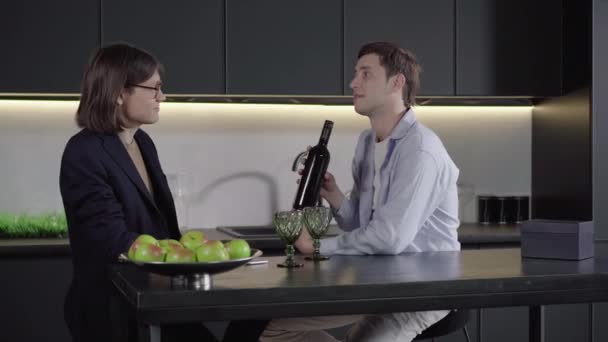 Side view portrait of young handsome man holding wine bottle and talking with boyfriend. Brunette Caucasian gay couple celebrating happy anniversary indoors. Lgbt love, romance, lifestyle. - Video