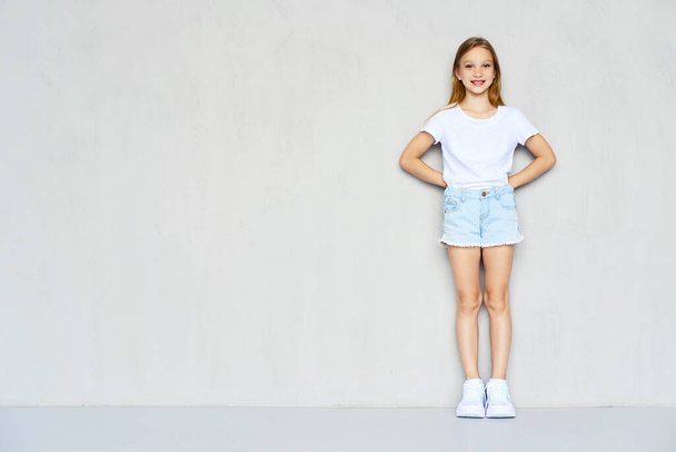 Young sportive girl in white t-shirt and jeans shorts leaning to the wall with both hands behind her back - Φωτογραφία, εικόνα
