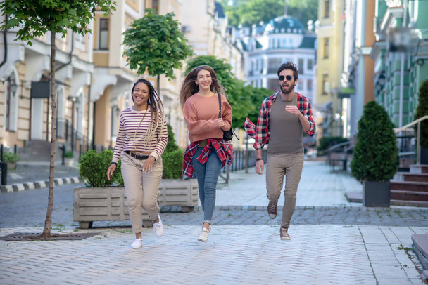 Young adult guy and two girls having fun running along street - Φωτογραφία, εικόνα