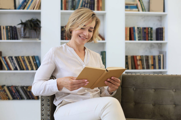 Attractive middle aged woman enjoying reading a book sitting on the sofa in her living room smiling while she reading - Foto, Imagen