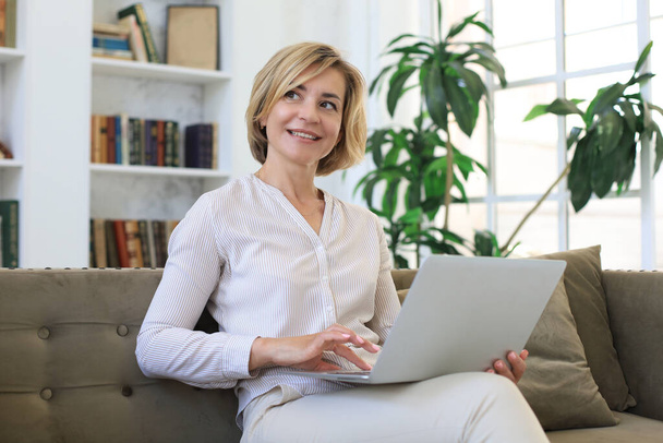 Cheerful middle aged woman using laptop while sitting on sofa at home - Foto, imagen