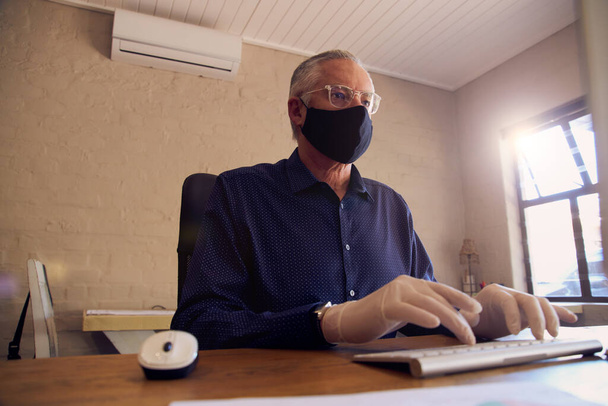 コンピュータの前にフェイスマスクを着用してデスクで働くビジネスマン. - 写真・画像