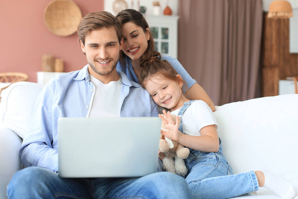 Positive friendly young parents with smiling little daughter sitting on sofa together answering video call on laptop and waving hand in greeting while relaxing at home on weekend - Zdjęcie, obraz