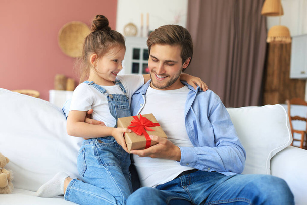 Cute kid daughter make surprise to daddy, little girl presents gift box to father sit on sofa. Father's day - Fotografie, Obrázek