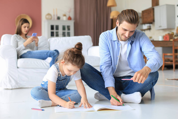 Happy father smilling daughter lying on warm floor enjoying creative activity, drawing pencils coloring pictures in albums, mother resting on couch, family spend free time together - Фото, изображение