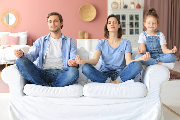 Calm young family with little daughter sit on couch practice yoga together, happy parents with small preschooler girl child rest on sofa meditate relieve negative emotions on weekend at home - Photo, image