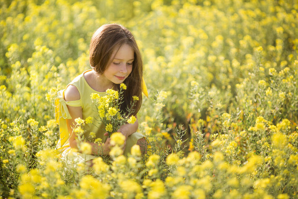 teenage girl in a yellow dress picks flowers in a yellow field in summer - Фото, изображение